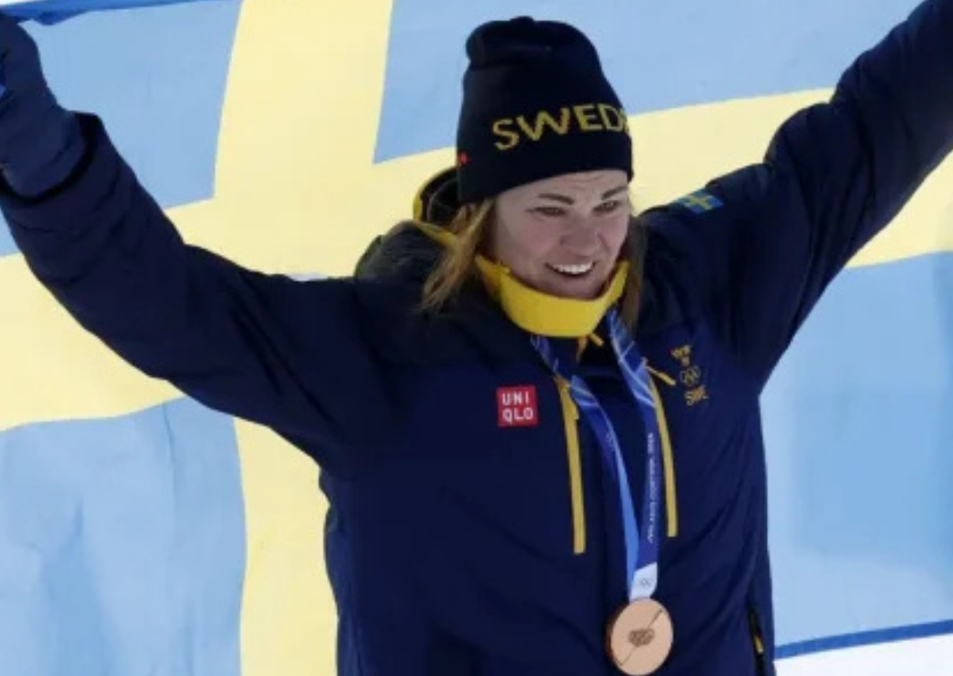 Anna Swenn Larsson wearing a navy blue longline puffer coat with Swedish Olympic crest and yellow zippers at the Milano Cortina 2026 Winter Olympics medal ceremony