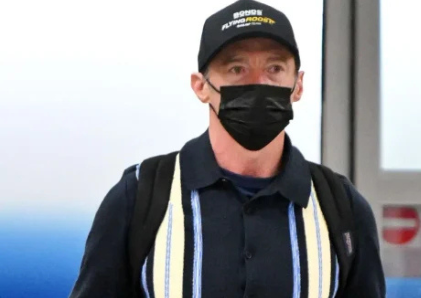 Hugh Jackman wearing a navy blue striped cardigan-style shirt with matching trousers, black cap, face mask, backpack, and suitcase at the airport
