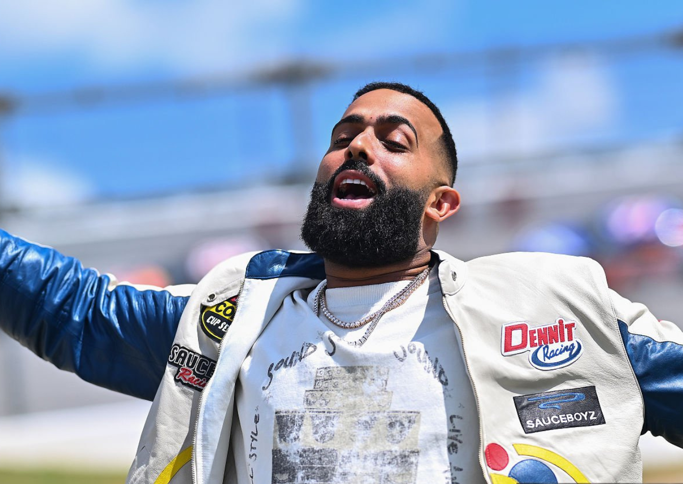 Eladio Carrion wearing a cream and blue racing leather jacket with sponsor patches, distressed jeans, and silver chains during NASCAR Cup Series Jack Link’s 500 at Talladega Superspeedway