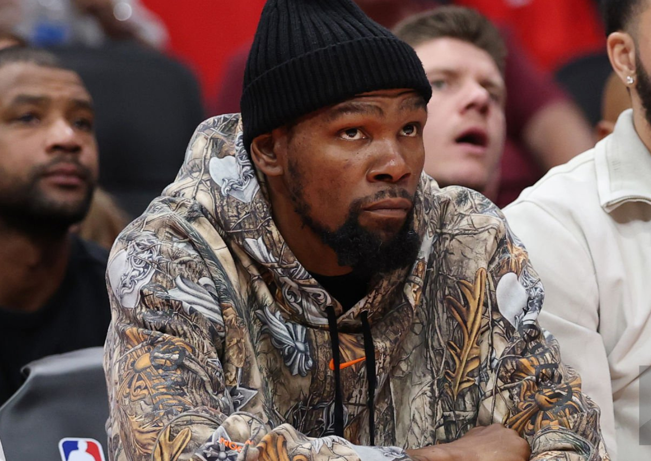 Kevin Durant wearing tan and brown RealTree camo hoodie with Chrome Hearts graphics, black cargo sweatpants, beanie, and Nike sneakers during 2026 NBA Playoffs game