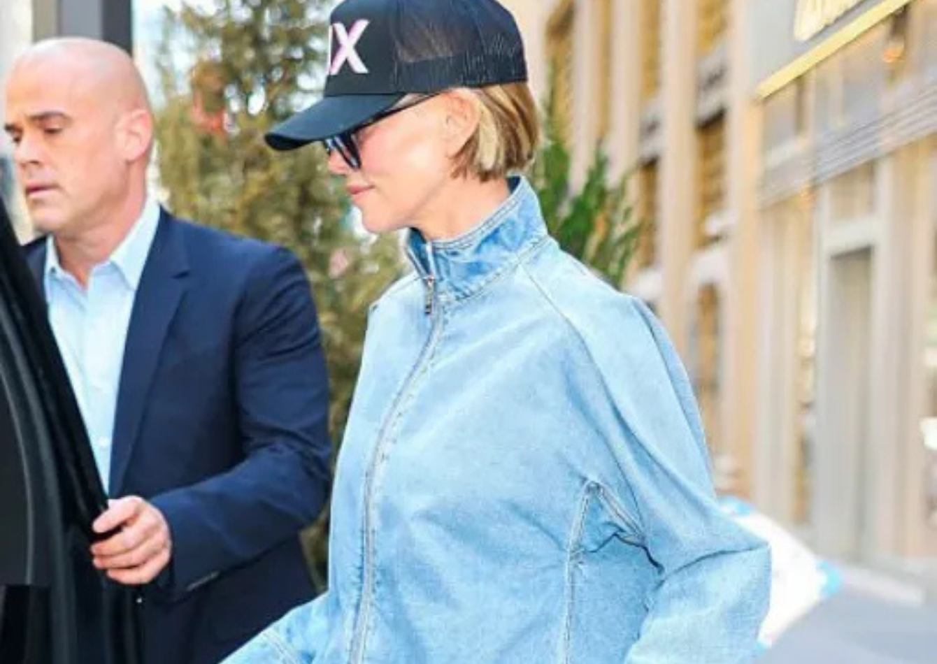 Charlize Theron wearing a light-wash oversized denim jacket with wide-leg white trousers, black cap, sunglasses, and white sneakers in NYC 2026 street style