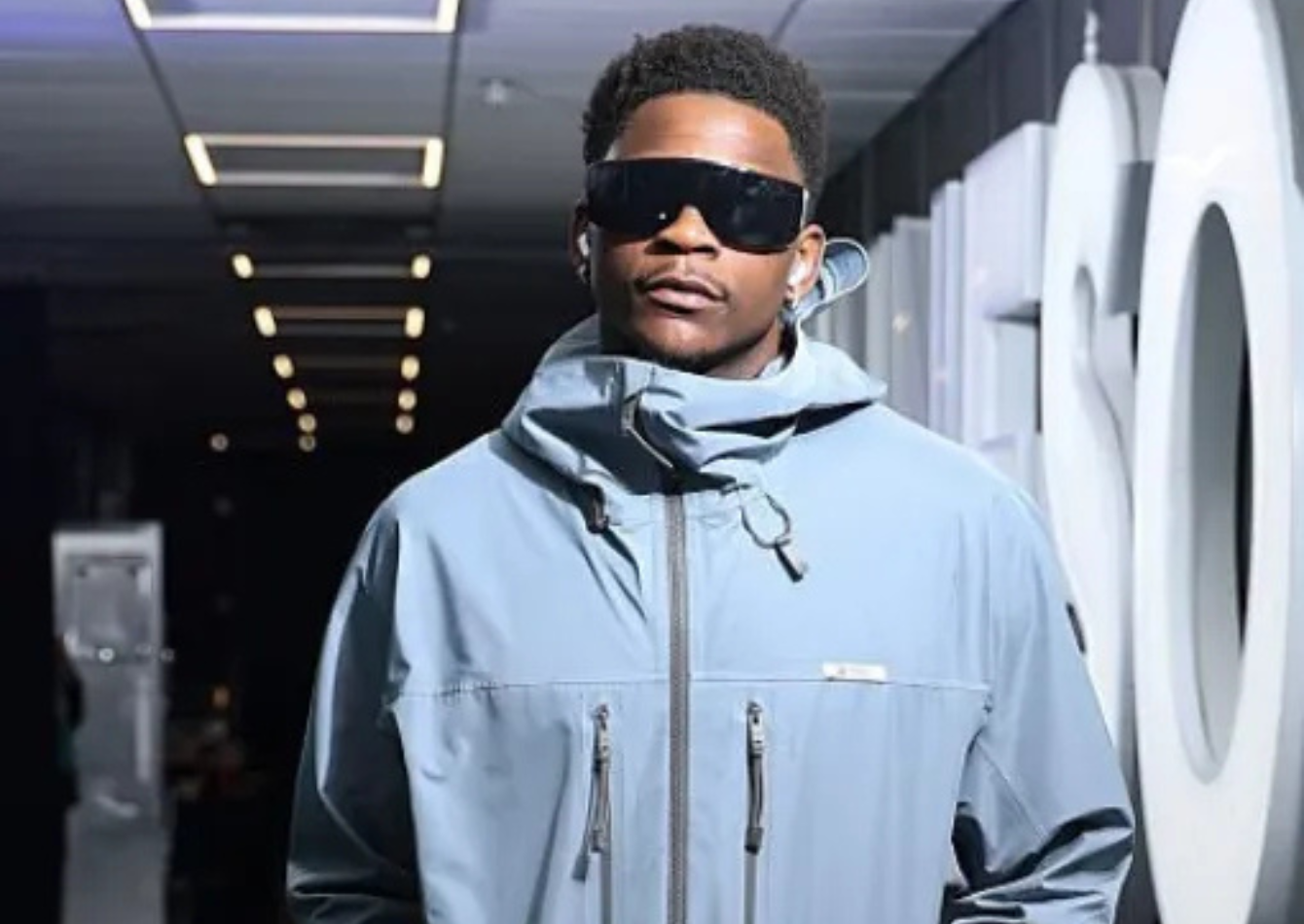 Anthony Edwards wears a light blue technical hooded jacket with dark green cargo pants and sunglasses, creating a bold modern streetwear look in a stadium corridor.