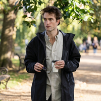 Robert Pattinson wearing a black parachute-fabric hooded technical jacket on the set of The Drama (2026), styled with a gray Oxford shirt, dark-rimmed glasses, and minimal accessories, showcasing a modern techwear-inspired cinematic look.