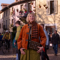 Kerri Kenney wearing a burnt-orange faux fur reversible jacket with black leather trim from The Four Seasons Season 2, styled with an A-line skirt, patterned scarf, and tartan bag in a cinematic outfit scene.