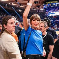 Taylor Swift courtside at NBA Finals wearing a royal blue cropped “Stevie Knicks” graphic tee with denim jeans and streetwear styling at Madison Square Garden.