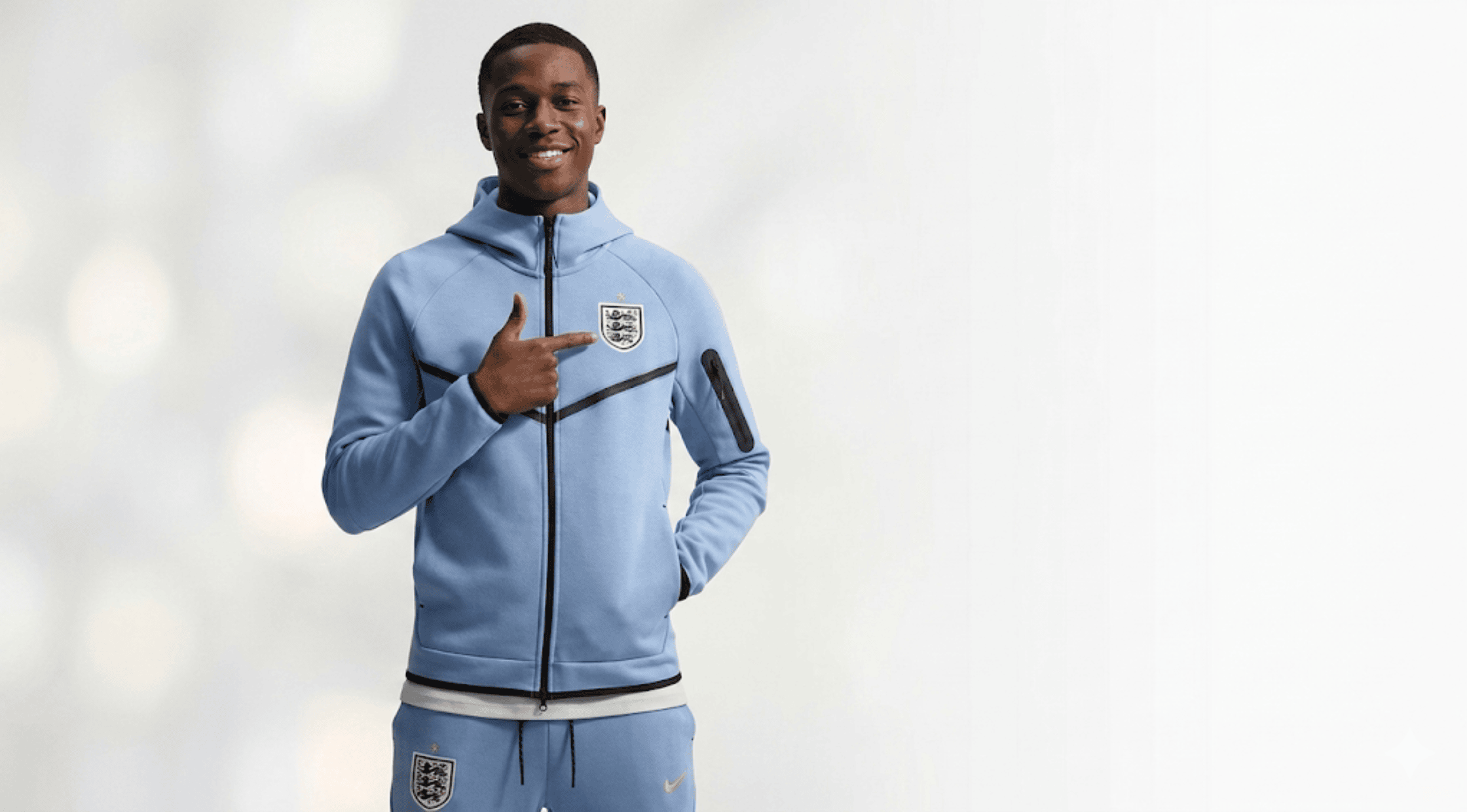 A man smiling and pointing at the official England Three Lions crest on his light blue tech fleece windrunner hoodie jacket.