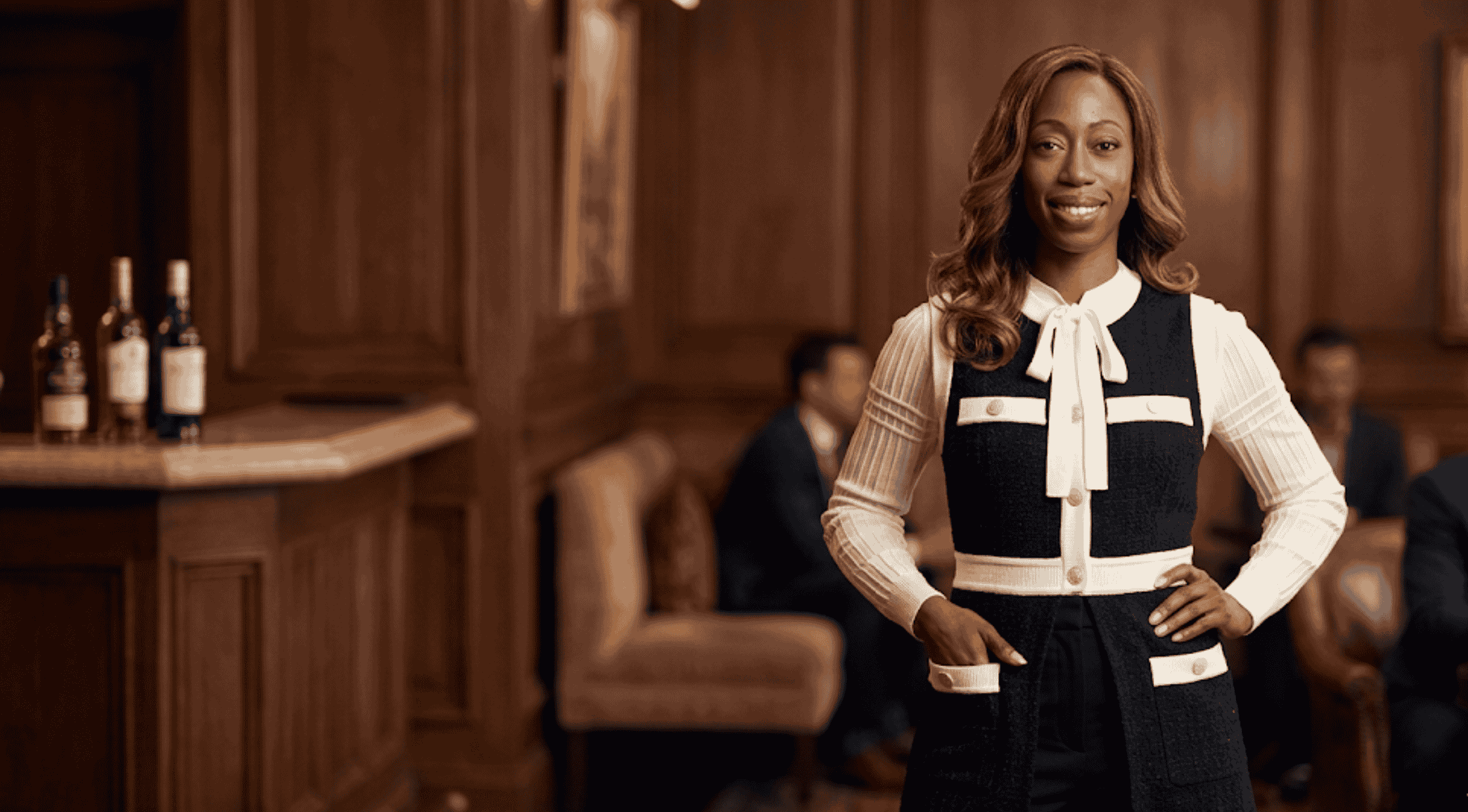 Nikki Amuka-Bird smiling in a black and white Sandro sleeveless cardigan while standing inside a warmly lit lounge.