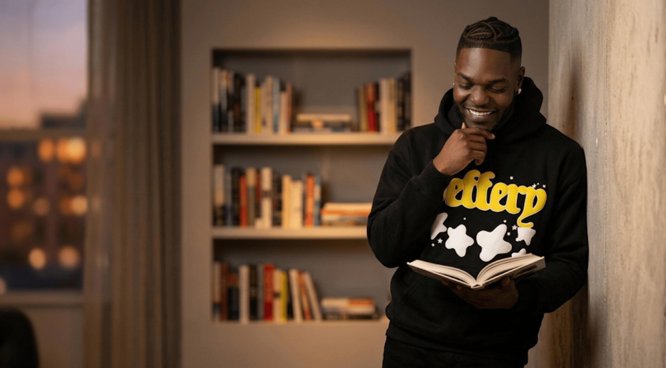 A man leaning against a concrete wall wearing a black hoodie with yellow "Jeffery" cursive text and abstract white stars while reading a book.